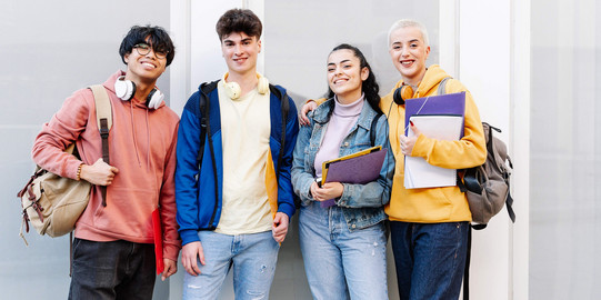 Vier internationale Studierende mit Mappen, Rucksäcken und Kopfhörern vor einem weißen Gebäude